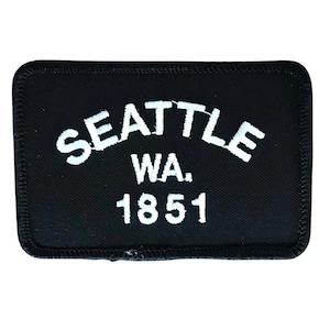 Pode incluir: Etiqueta bordada preta com texto branco que diz "SEATTLE WA. 1851".