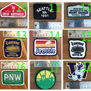 May include: A collection of eight embroidered patches featuring various designs related to Seattle, Washington. The patches are in different colours, including red, white, blue, green, yellow, and black. The patches feature text such as "The Great Northwest", "Seattle, WA", "Mount Rainier National Park", "Kingdome", "PNW", and "Seattle".