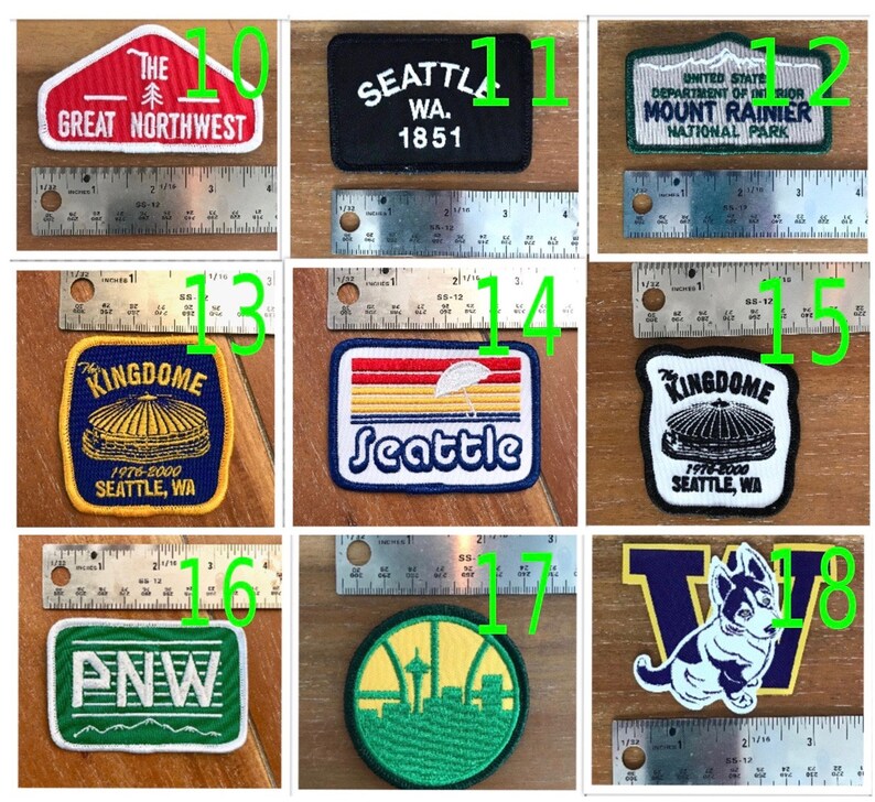 May include: A collection of eight embroidered patches featuring various designs related to Seattle, Washington. The patches are in different colours, including red, white, blue, green, yellow, and black. The patches feature text such as "The Great Northwest", "Seattle, WA", "Mount Rainier National Park", "Kingdome", "PNW", and "Seattle".