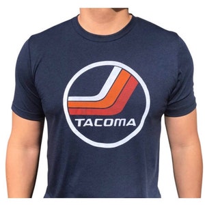 May include: A navy blue t-shirt with a white circle graphic featuring a red and orange stripe design and the word "TACOMA" in white text.
