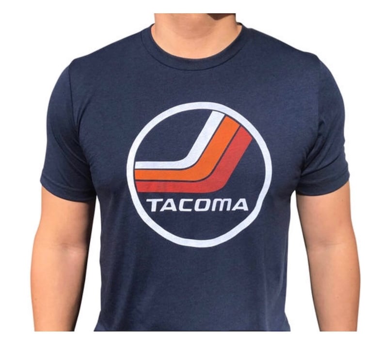 May include: A navy blue t-shirt with a white circle graphic featuring a red and orange stripe design and the word "TACOMA" in white text.