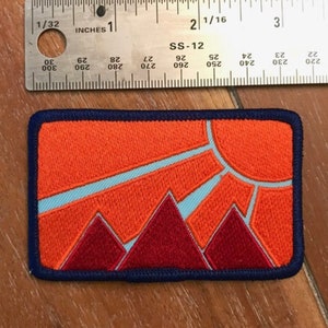 May include: An orange, red, and blue embroidered patch with a sun and mountains. The patch has a navy blue border and the text "SS-12" is printed on the top.