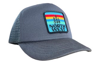 May include: Gray trucker hat with a square patch featuring a colorful mountain and tree design. The patch has the words "NORTH WEST" in white block letters. The hat has a curved brim and mesh back.