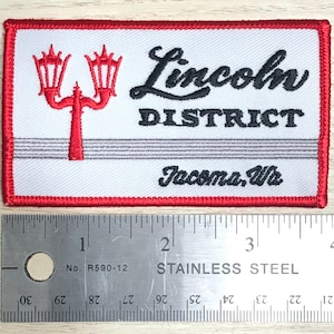 May include: Embroidered patch with a white background, red border, and the words "Lincoln DISTRICT Tacoma, Wa" in black script. A red lamp post design is on the left. The patch is rectangular and is about 10 cm wide.