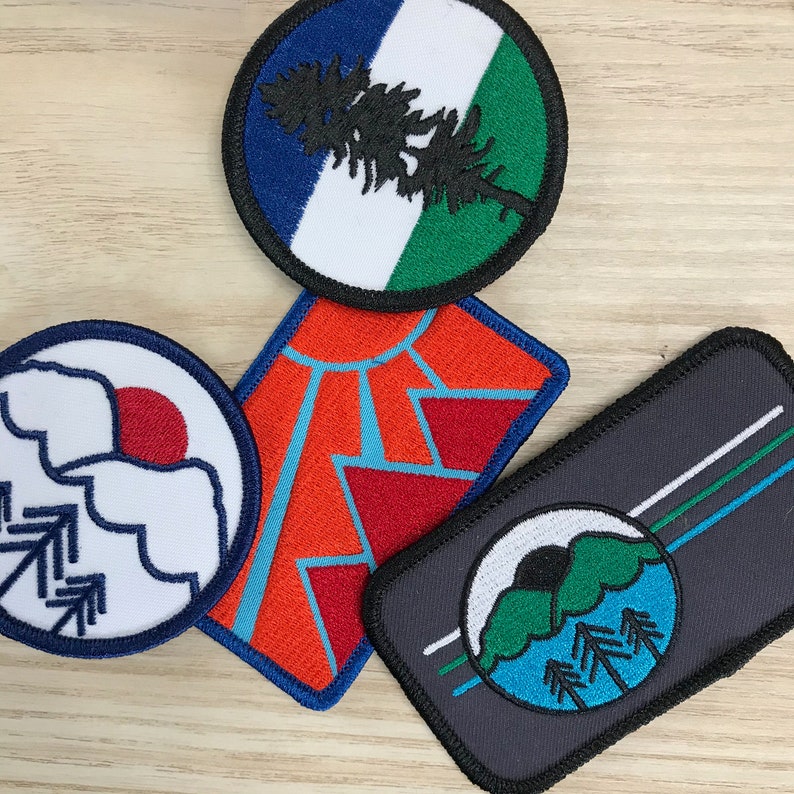 May include: Four embroidered iron-on patches with different designs. One patch features a blue, white and green design with a pine tree. Another patch features a red, white and blue design with a mountain range. A third patch features a white and blue design with a mountain and trees. The final patch features a black, white and green design with a mountain and trees.
