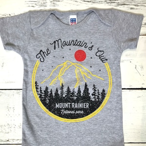 May include: Gray baby onesie with a yellow and red graphic of a mountain range with the text "The Mountain's Out" and "Mount Rainier National Park".