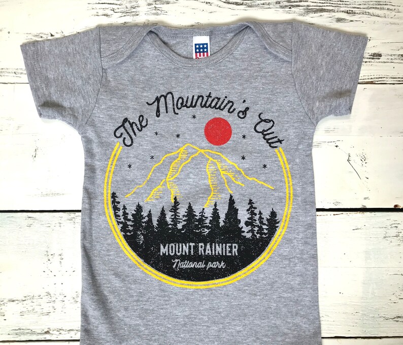 May include: Gray baby onesie with a yellow and red graphic of a mountain range with the text "The Mountain's Out" and "Mount Rainier National Park".