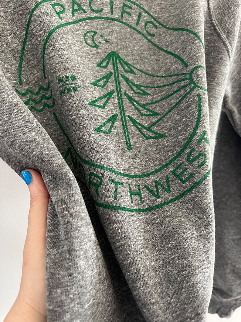 May include: Gray sweatshirt with a green circular graphic design featuring a stylized pine tree, a crescent moon, and the text "PACIFIC NORTHWEST".