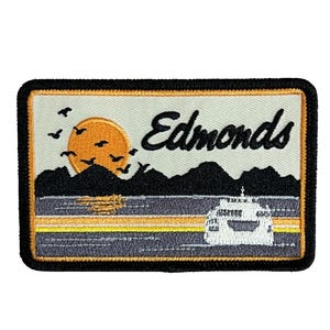 May include: Embroidered patch featuring a sunset over mountains, birds, and a ferry. The word "Edmonds" is in black script. The patch has a black border and a cream-colored background.