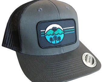 May include: A gray trucker hat with a black mesh back. The hat features a black and blue embroidered patch with a mountain scene and a white circle with blue and green accents.