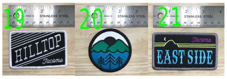 May include: Three embroidered patches featuring black, white, blue and green thread. The first patch is black with white stripes and the text "HILLTOP Tacoma". The second patch is blue with a white background and a green mountain scene. The third patch is black with a yellow crescent moon and the text "EAST SIDE Tacoma" in blue.