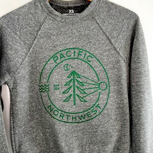 May include: Gray sweatshirt with a green circle logo that says "PACIFIC NORTHWEST" with a stylized pine tree, moon, and waves inside the circle. The sweatshirt has a size tag that says "XS SWEETPEA CO. BOY."