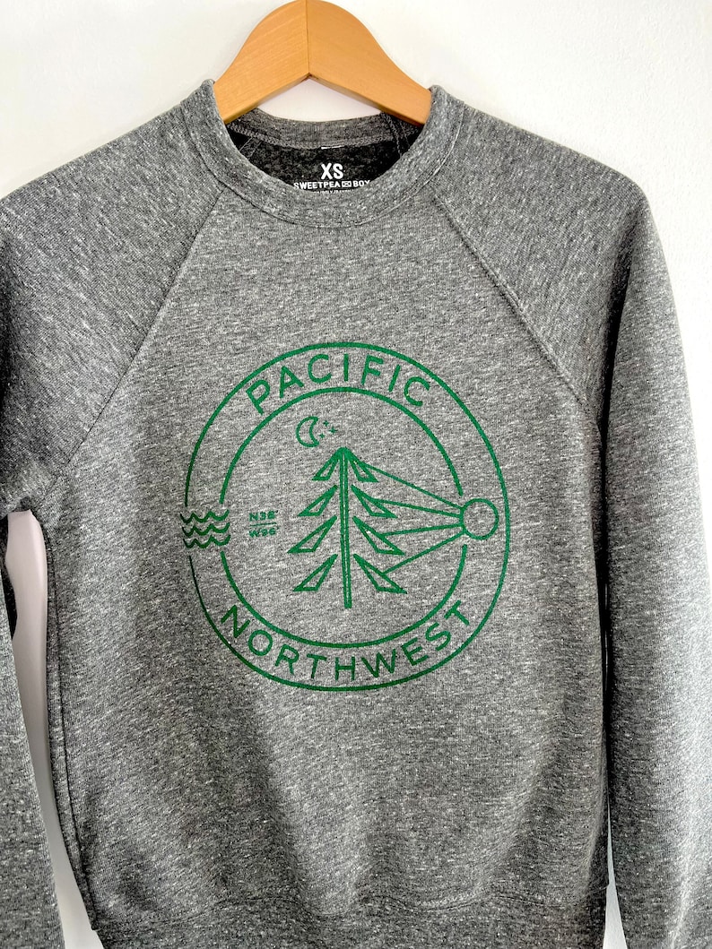 May include: Gray sweatshirt with a green circle logo that says "PACIFIC NORTHWEST" with a stylized pine tree, moon, and waves inside the circle. The sweatshirt has a size tag that says "XS SWEETPEA CO. BOY."