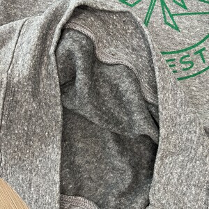 May include: A close-up of the inside of a gray heather t-shirt with a green graphic of a stylized pine tree and the text "EST."