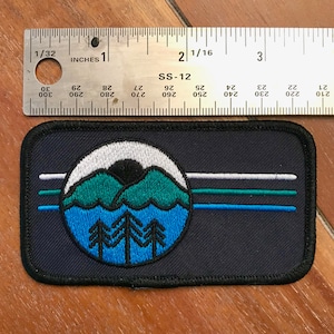 May include: A blue, green and white embroidered patch featuring a mountain scene and three trees. The patch is on a black background and has the text "SS-12" above the scene.