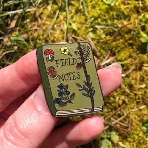 May include: Green enamel pin shaped like a book with the words "Field Notes" on the cover. The pin is decorated with various nature-themed illustrations, including a mushroom, a flower, a leaf, an acorn, and a pine branch.
