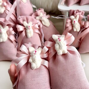 May include: Pink burlap favor bags with white bunny appliques and pink satin ribbon ties. The bags are arranged in a display on a white shelf.