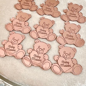 May include: A collection of rose gold mirror acrylic bear favors with the text "Baby Malani" engraved on each bear.