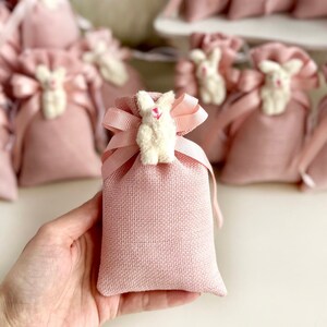 May include: Pink burlap favor bags with white bunny charms. The bags are tied with a pink ribbon and are perfect for a baby shower or birthday party.
