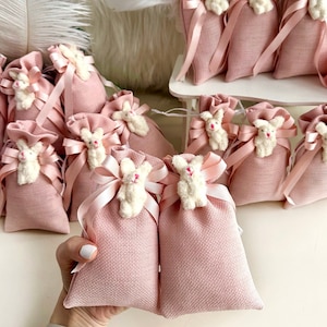 May include: Pink fabric gift bags with white bunny appliques and pink ribbon ties. The bags are arranged in a group on a white surface.