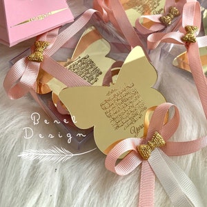 Pode incluir: Caixas de presente rosa e brancas com espelhos dourados em forma de borboleta. Cada espelho tem uma inscrição dourada e o nome "Yara" está escrito em um dos espelhos.