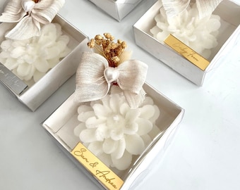 May include: White flower-shaped candles in small white boxes with a clear top. Each box has a small bouquet of dried flowers and a gold tag with text. The text on the tags reads "Baby from" and "Evan & Andrea".