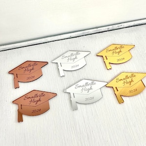 Puede incluir: Cinco gorros de graduación hechos de acrílico espejado en oro, plata y cobre. Cada gorro tiene el texto "Smallville High 2024" grabado.