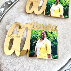 May include: Two gold mirror acrylic plaques with the number 60 and the name Linda. The plaques have a photo of a person in a yellow jacket. The text on the plaques reads "July 15, 2022".