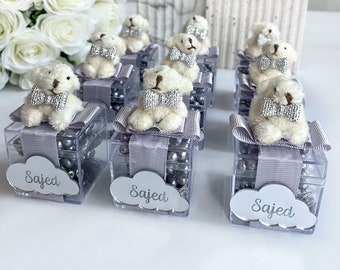 May include: Nine clear favor boxes with silver lids and a white teddy bear with a silver bow tie sitting on top of each box. Each box is filled with silver beads and has a silver cloud with the name "Sajed" printed on it.