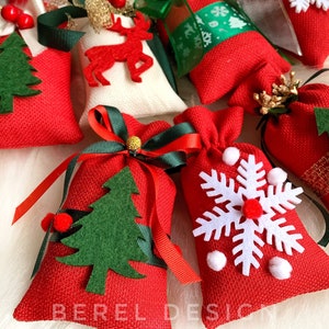 Puede incluir: Una colecci&oacute;n de peque&ntilde;as bolsas de regalo de tela roja con varias decoraciones con tem&aacute;tica navide&ntilde;a, que incluyen un reno, un copo de nieve, un &aacute;rbol de Navidad y una cinta. Las bolsas est&aacute;n llenas de peque&ntilde;os regalos y son perfectas para un calendario de adviento navide&ntilde;o.