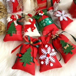 Puede incluir: Surtido de bolsas de regalo de tela de Navidad rojas y blancas con varios adornos de fieltro, incluyendo copos de nieve, renos y &aacute;rboles de Navidad. Las bolsas est&aacute;n atadas con cinta verde, roja y dorada.