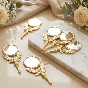 May include: A collection of gold-colored handheld mirrors, each featuring a circular glass surface attached to a handle shaped like a winged figure. The mirrors are arranged on a white marble slab and a smooth surface, with some placed individually and others grouped together.