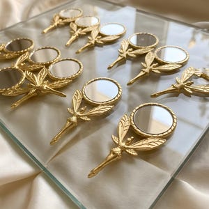 May include: A collection of small, gold-toned metal hand mirrors, each featuring a circular reflective surface framed by a decorative handle shaped like a winged insect. The mirrors are arranged in a grid pattern on a clear glass surface.