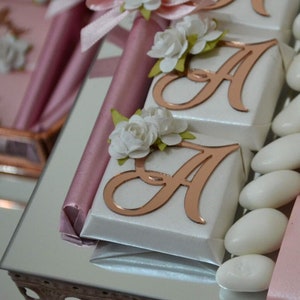 May include: A tray of white and pink favors with a rose gold letter A on each. The favors are decorated with white flowers and pink ribbon.