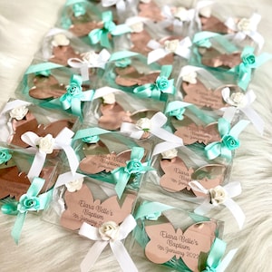 Puede incluir: Una colecci&oacute;n de peque&ntilde;as cajas transparentes que contienen espejos en forma de mariposa de oro rosa. Cada caja est&aacute; decorada con una cinta blanca y azul claro, una flor blanca y un mensaje personalizado que dice "Bautizo de Elara Belle 9 de enero de 2022".