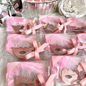 May include: Pink favour boxes with clear lids filled with small rose gold masquerade masks. Each mask has a white feather and a pink ribbon tied around it. The masks are personalised with the name "Sienna's" and the date "May 6, 2023".