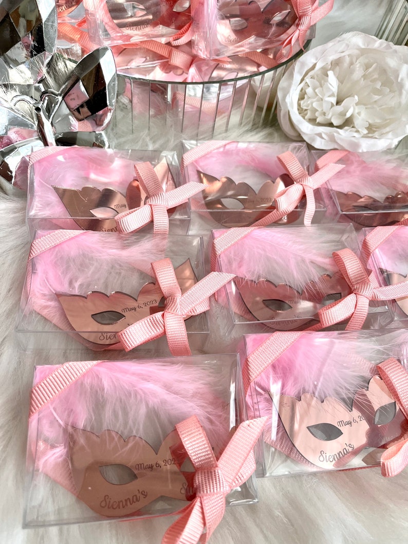 May include: Pink favour boxes with clear lids filled with small rose gold masquerade masks. Each mask has a white feather and a pink ribbon tied around it. The masks are personalised with the name "Sienna's" and the date "May 6, 2023".