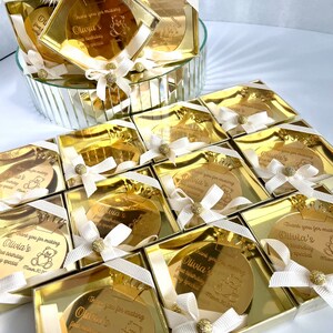 May include: Gold favor boxes with a round, gold mirror with a crown and a teddy bear design. The mirror has the text "Thank you for making Olivia's first birthday very special March 30, 2023".