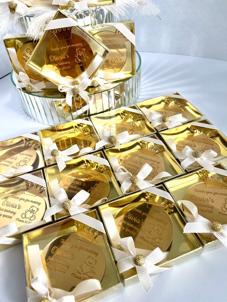 May include: Gold favor boxes with a round, gold mirror with a crown and a teddy bear design. The mirror has the text "Thank you for making Olivia's first birthday very special March 30, 2023".