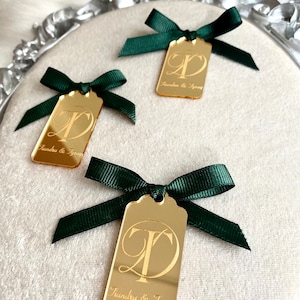 May include: Three gold mirror acrylic tags with green ribbon bows. Each tag has a cursive letter 'T' and 'D' intertwined and the text "Tiandra & Tyray" below.