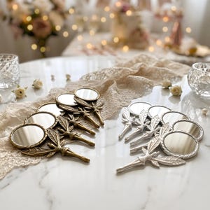 May include: A collection of small handheld mirrors featuring fairy-shaped handles with detailed wings. The set includes several mirrors with a metallic gold finish and several mirrors with a metallic silver finish, all arranged on a white surface.
