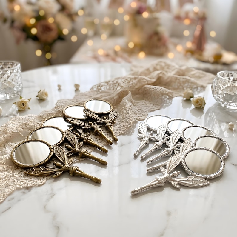 May include: A collection of small handheld mirrors featuring fairy-shaped handles with detailed wings. The set includes several mirrors with a metallic gold finish and several mirrors with a metallic silver finish, all arranged on a white surface.