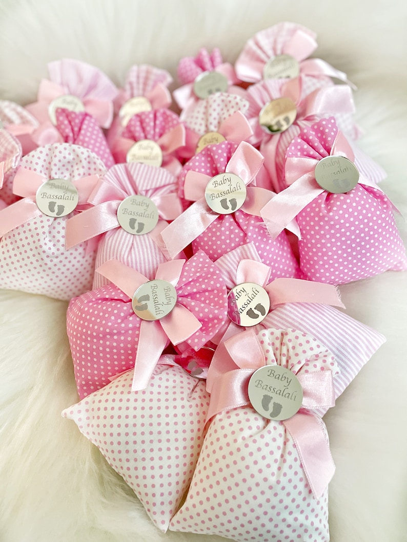 May include: Pink and white fabric gift bags with a silver circle tag that says "Baby Bassalali" in a cursive font. The bags are tied with a pink ribbon.