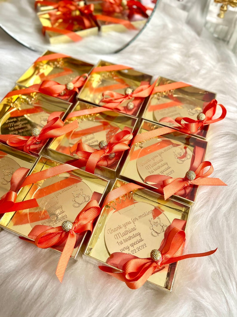 May include: Gold favor boxes with clear lids and orange ribbons. Each box contains a round, gold-colored, personalized acrylic ornament with a bear design and the text "Thank you for making Mathias' 1st birthday very special 22.2022".