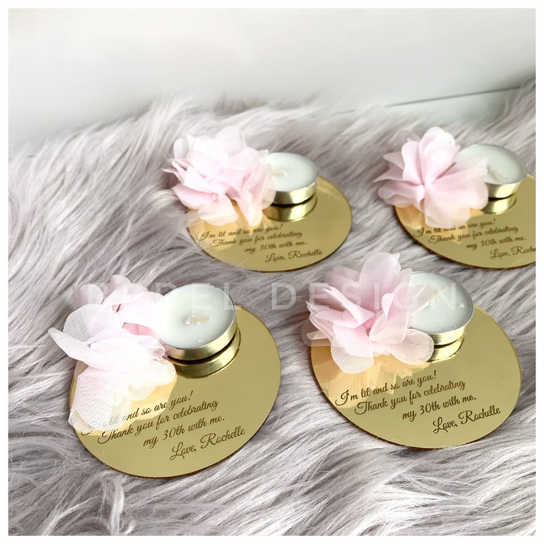 May include: Four gold round mirrored candle holders with a pink flower on top and a small white tea light candle. The holders have a personalized message engraved on them that says "I'm lit and so are you! Thank you for celebrating my 30th with me. Love, Rochelle"