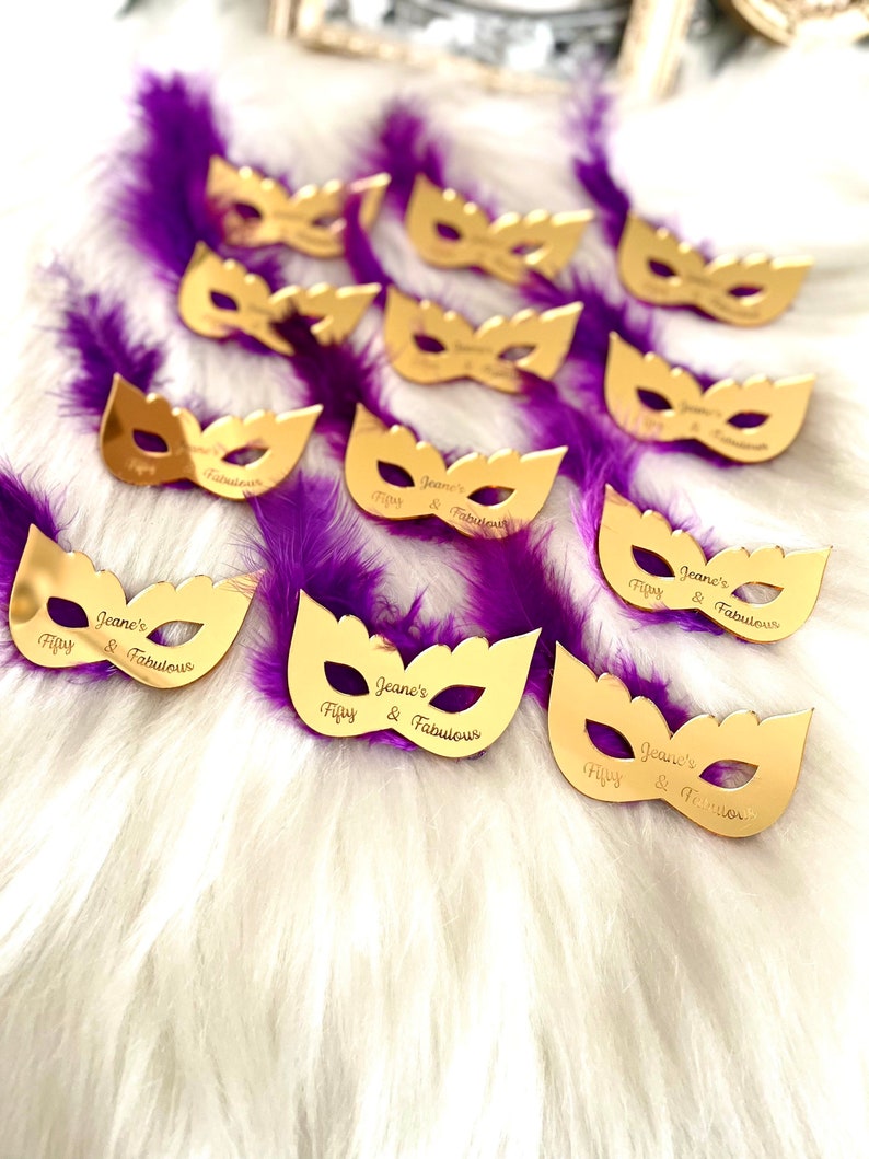 Puede incluir: Favores de fiesta en forma de m&aacute;scara de mascarada de color dorado con plumas moradas. Cada m&aacute;scara tiene el texto "Jeane's Fippy & Fabulous" escrito en ella.