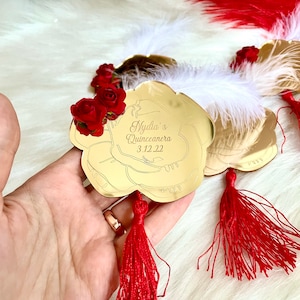 May include: Gold mirror with a red rose design and a red tassel. The mirror has the text "Nydia's Quinceanera 3.12.22" written on it.