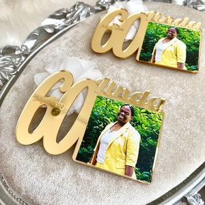 May include: Two gold mirror acrylic plaques with the number 60 and the name Linda. The plaques have a photo of a woman in a yellow suit. The text on the plaques reads "July 7, 2022".