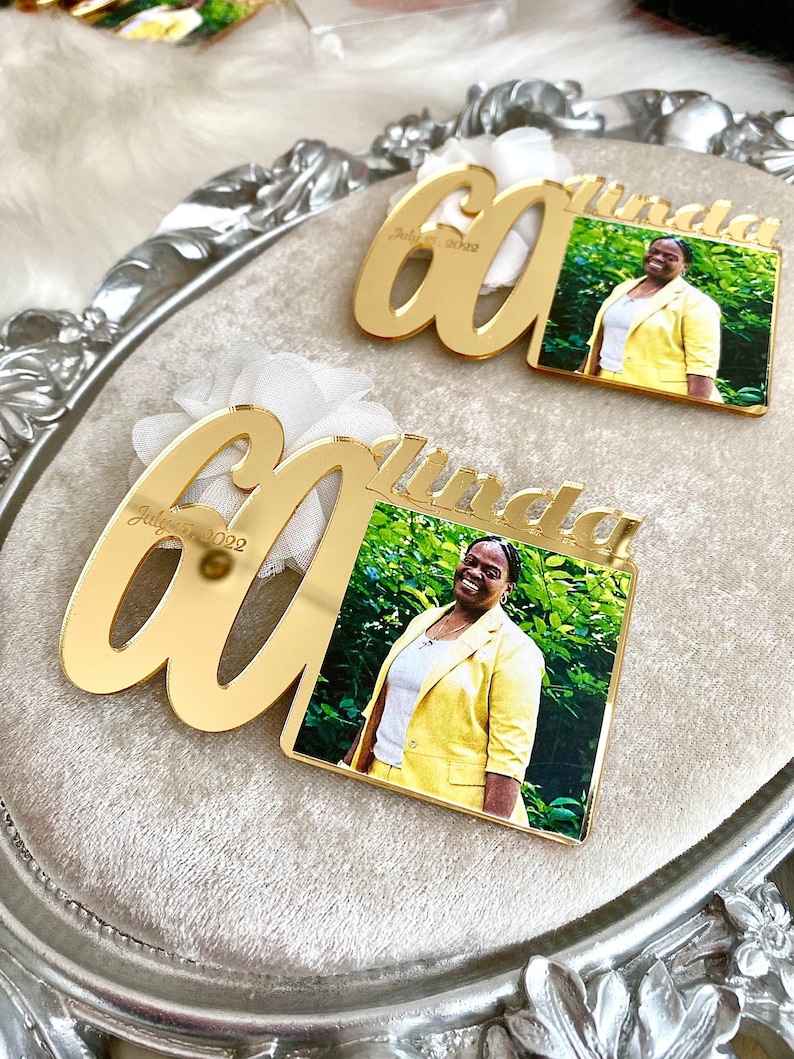 May include: Two gold mirror acrylic plaques with the number 60 and the name Linda. The plaques have a photo of a woman in a yellow suit. The text on the plaques reads "July 7, 2022".