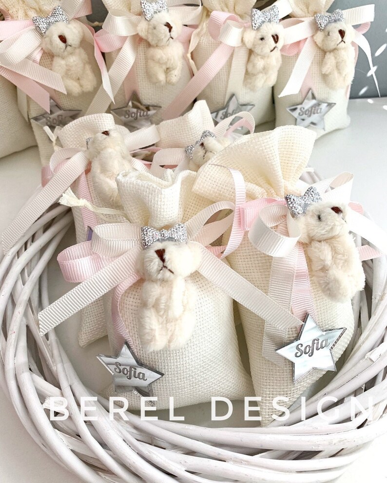 May include: White fabric gift bags with pink ribbon bows and a small white teddy bear with a silver crown. Each bag has a silver star with the name "Sofia" on it.